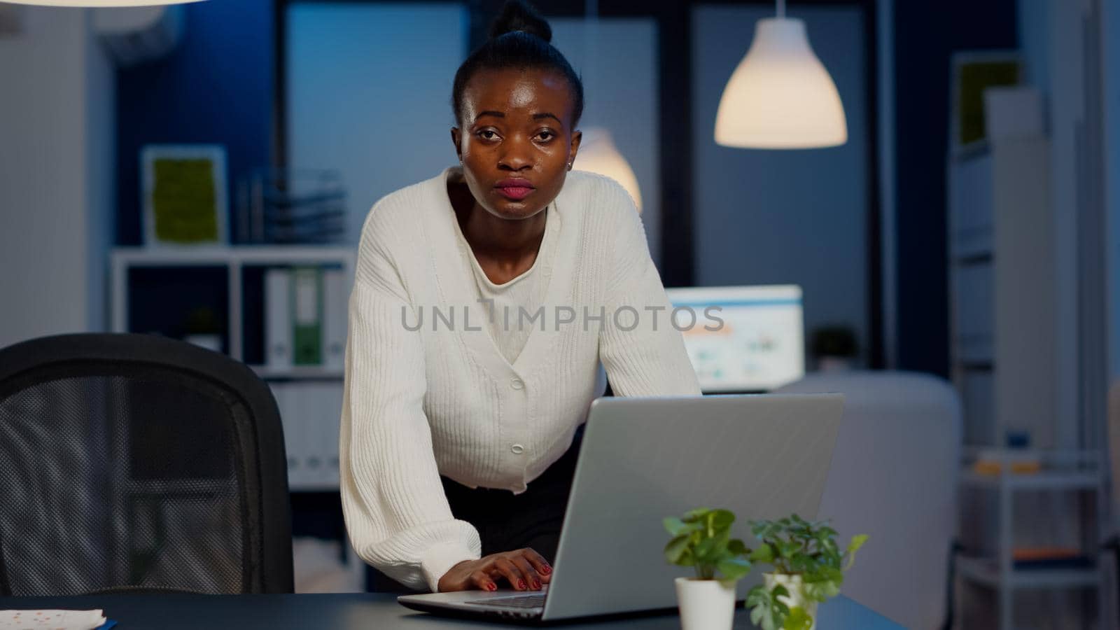 African business woman standing near desk looking at camera after reading mails on laptop working in start-up company late at night. Focused employee using technology network wireless doing overtime