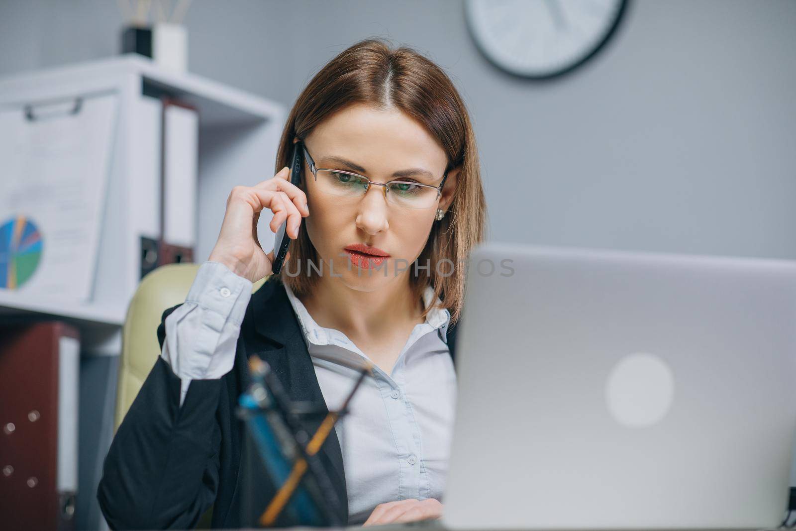 Young casual woman sitting at desk using laptop, answering call, talking on the phone, smiling teen speaking on mobile, having pleasant conversation on cell while working with computer in office