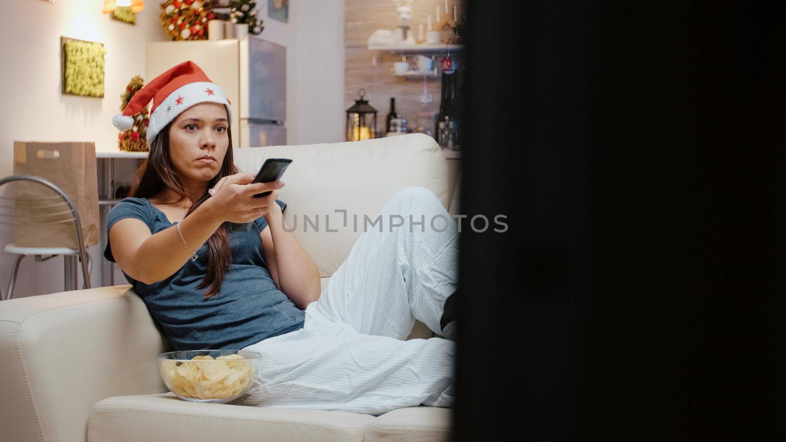 Unhappy person using TV remote control to switch channels on christmas festivity. Festive woman with santa hat watching television and eating chips from bowl to celebrate holiday