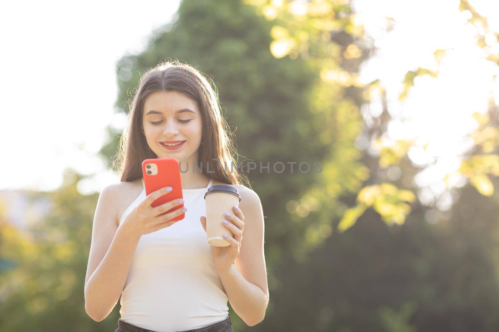 Attractive Girl Typing Messages on her Mobile. Portrait of Pretty Woman Standing in the Park with Smartphone and cup coffee in her Hands. Looking while typing Messages on her online Conversation by uflypro