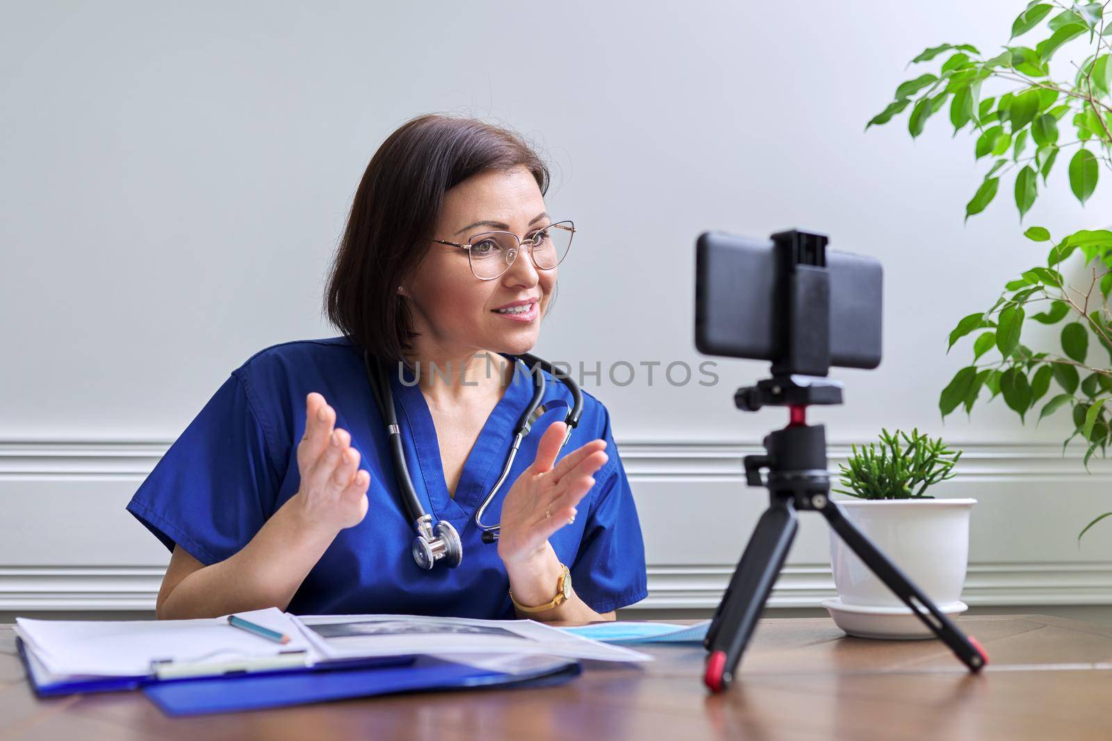 Female doctor with stethoscope consulting online using smartphone on tripod. Talking doctor looking at phone webcam, video meeting with patient. Medicine, technology, lifestyle, profession, people concept