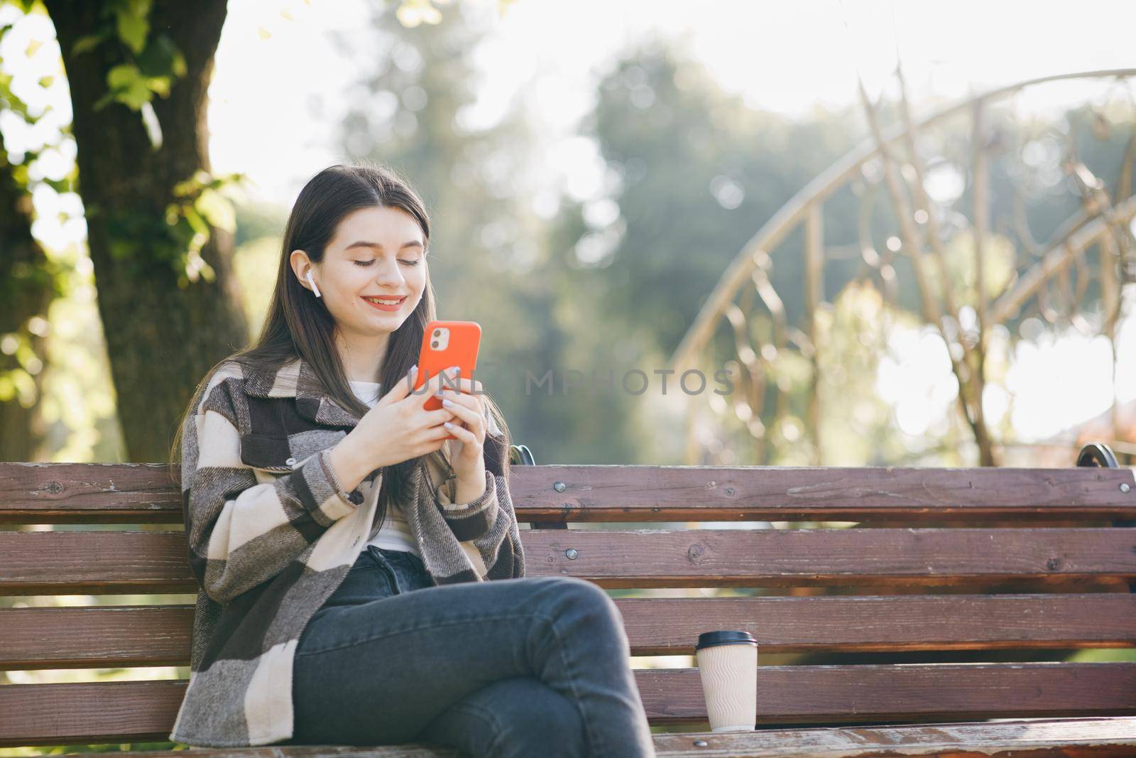 Portrait of girl wearing earphones and using smartphone listening music. Attractive woman browsing on mobile phone in park. City, urban background.