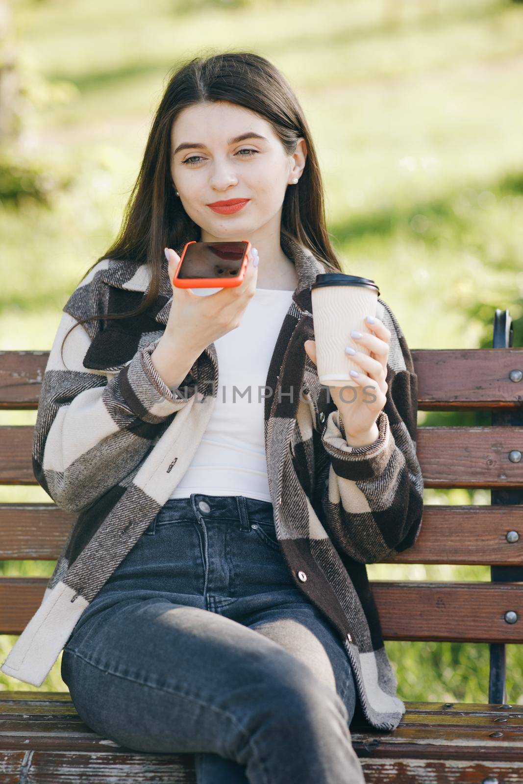 Girl using a smart phone voice recognition audio ai message speech function. Woman in trendy casual outfit sits on a bench in the city park by uflypro