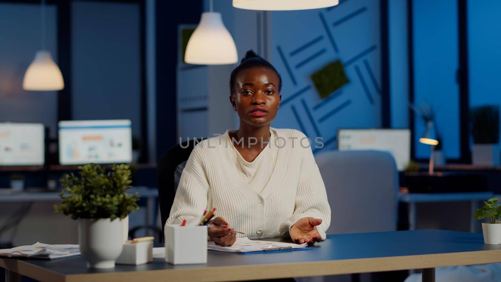 Pov of african businesswoman having video conference with team during midnight looking in camera in workplace. Freelancer using wireless technology network talking on virtual meeting doing overtime