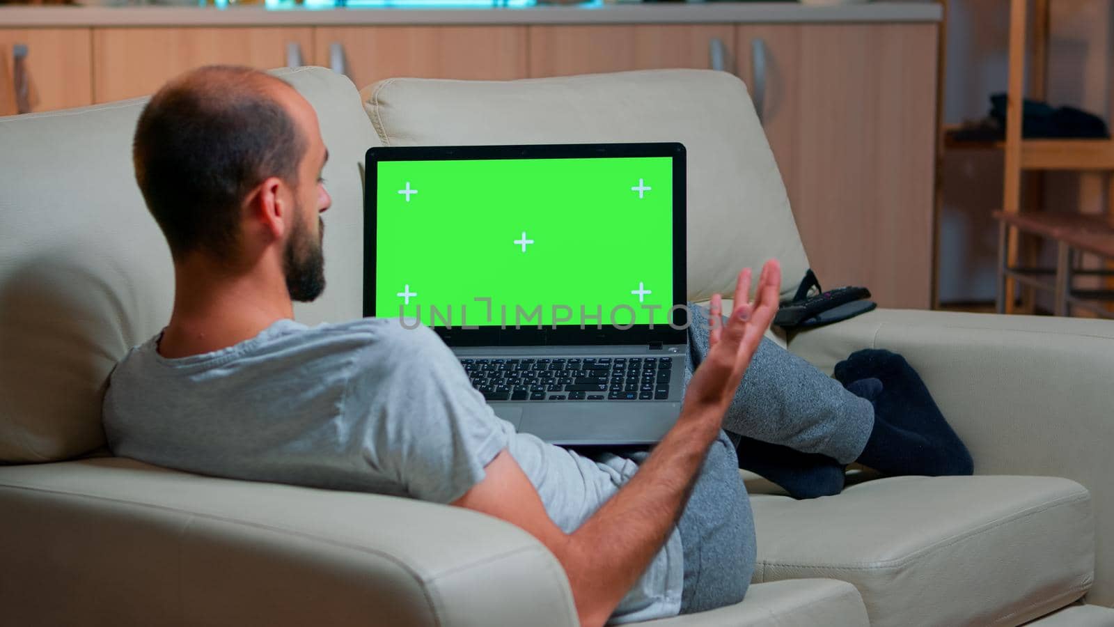 Focused man looking laptop computer with at mock up green screen chroma key display while laying on couch working at online e-learning project. Tired person using isolated pc late at night in kitchen
