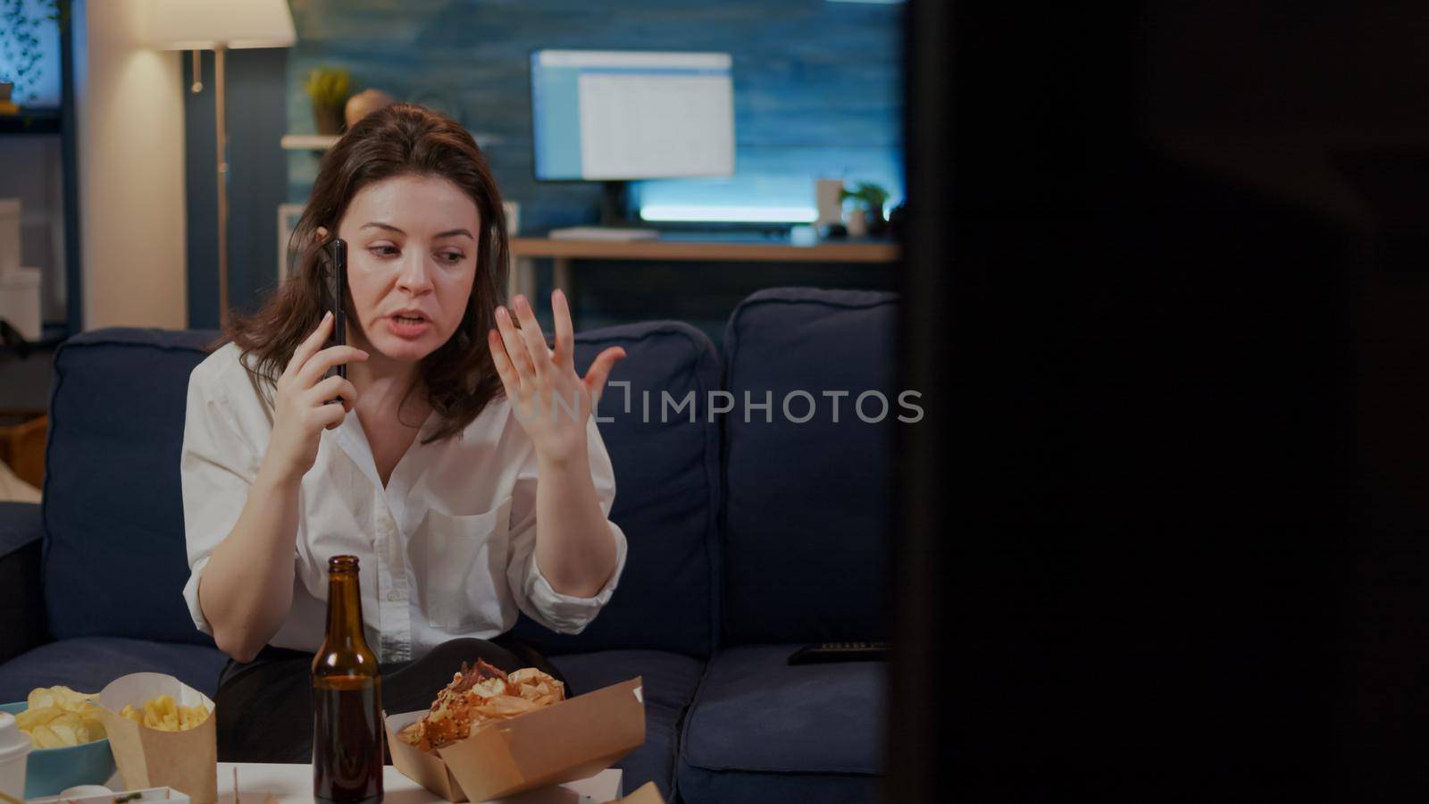 Annoyed woman talking on smartphone with boss after work at home while trying to eat takeaway food from delivery boxes. Person with fast food meal on table using phone for work call