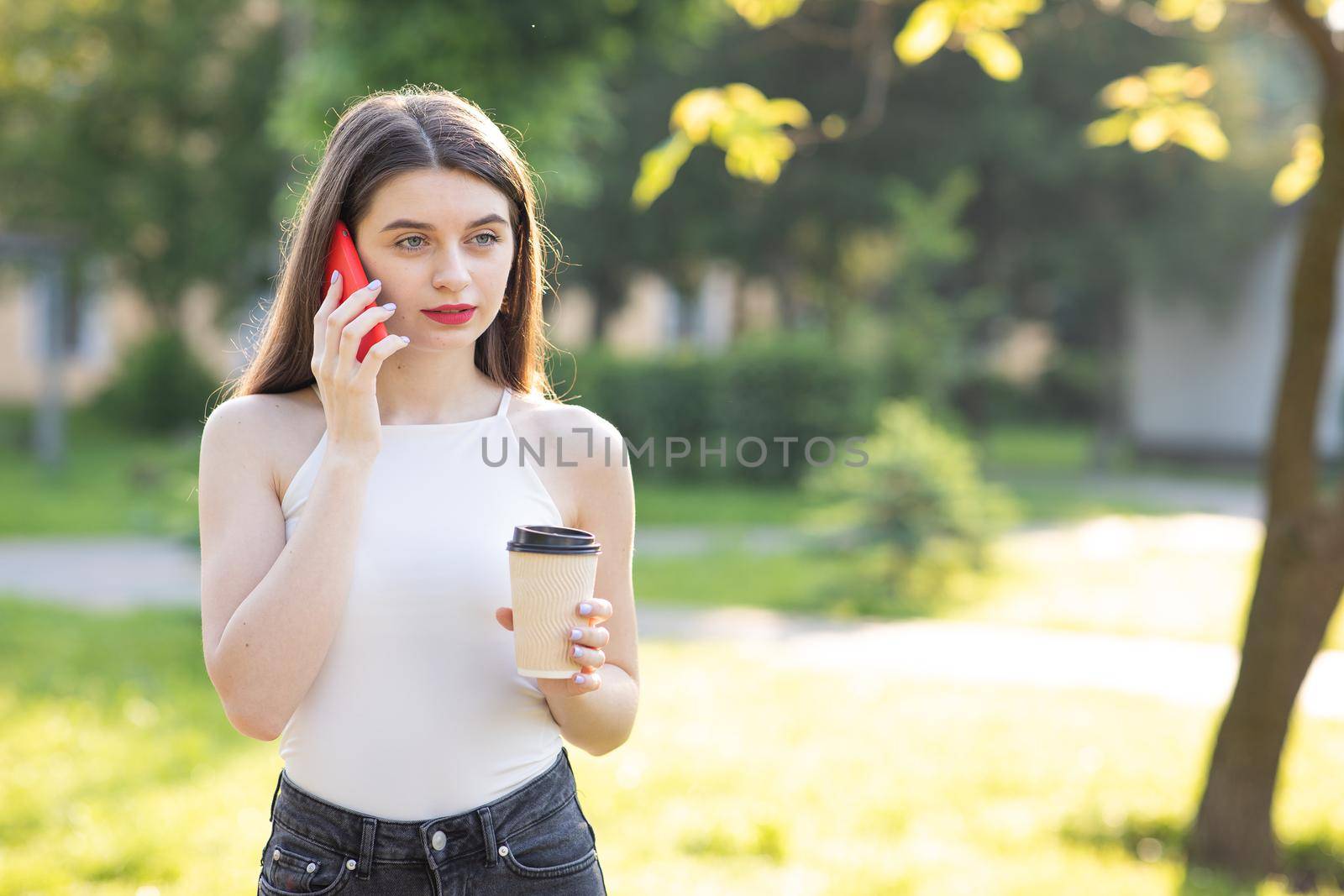 Young Attractive Girl Typing Messages on her Mobile. Portrait of Pretty Woman Standing in the Park with Smartphone and cup coffee in her Hands. Looking while typing Messages on her online Conversation