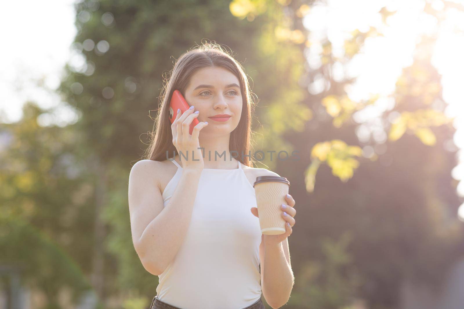 Portrait of Pretty Woman Standing in the Park with Smartphone and cup coffee in her Hands. by uflypro