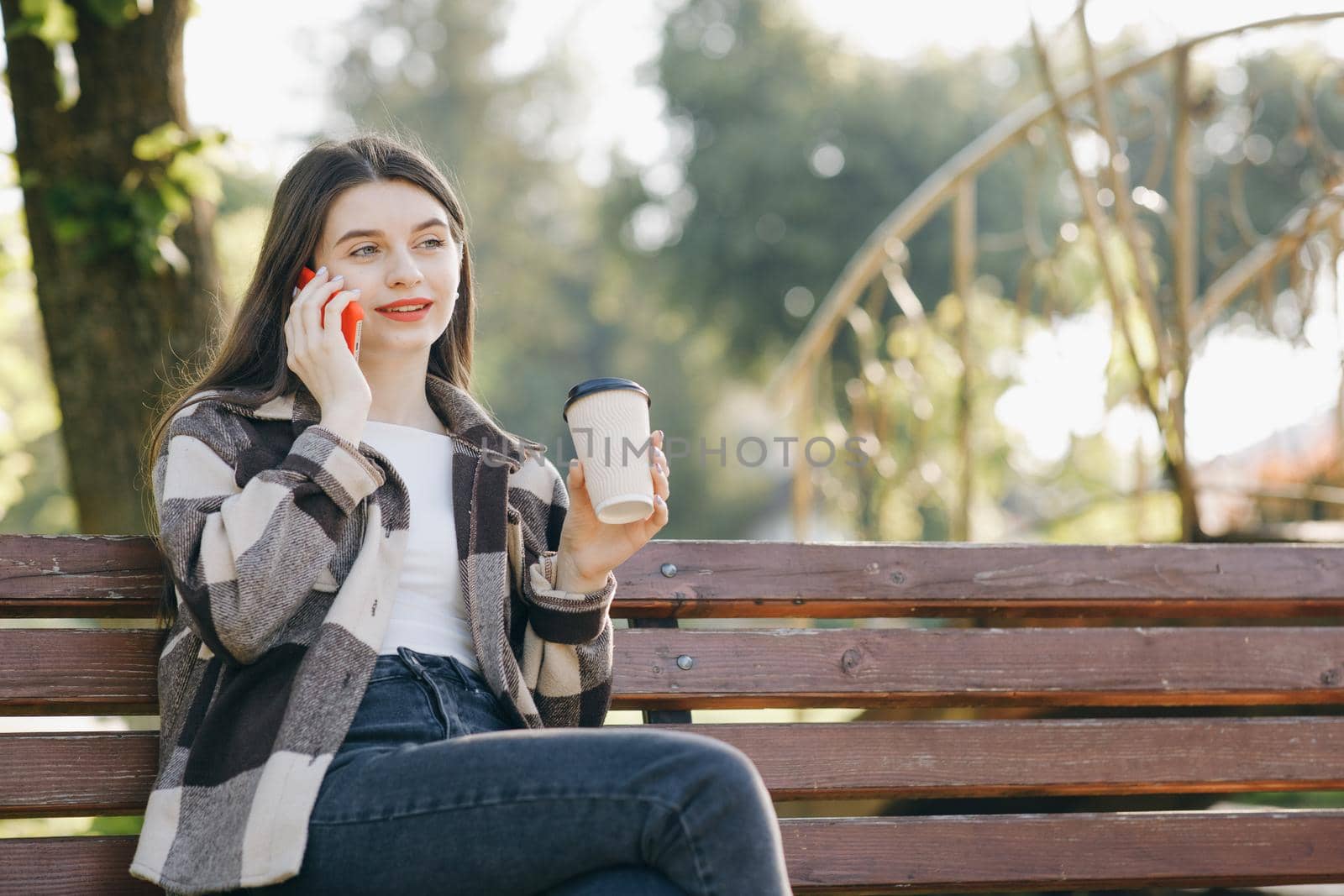 Portrait of a beautiful young girl talking on the phone sitting on a bench in the city park. Young woman dressed in stylish wear having phone conversation