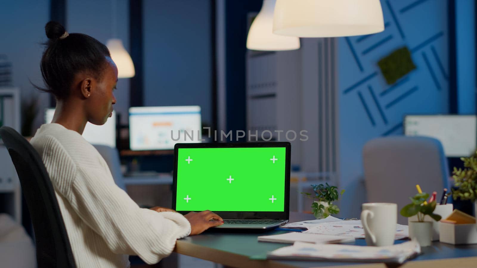 African manager typing at laptop with chroma key display during night time in start up business office working overtime. Freelancer watching mockup desktop monitor with green screen, writing datas
