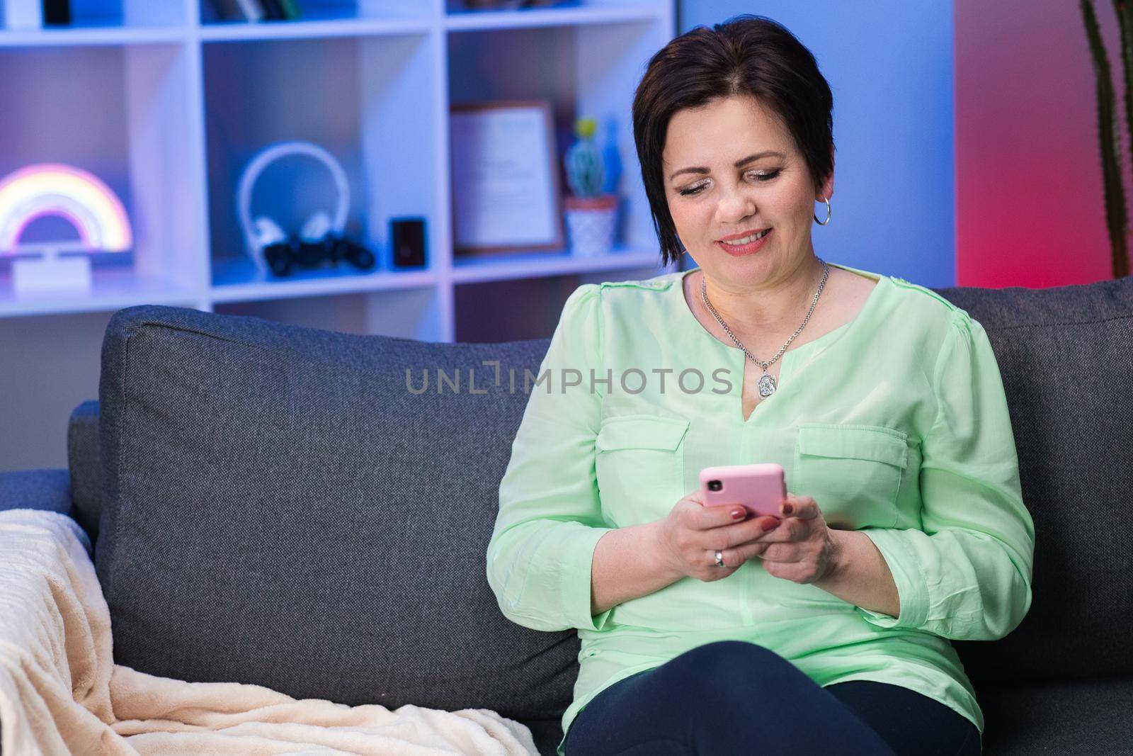 Caucasian Woman with Short Black Hair Sitting on Couch at Home, Holding Invisible Smartphone in Hand and Chatting with Someone