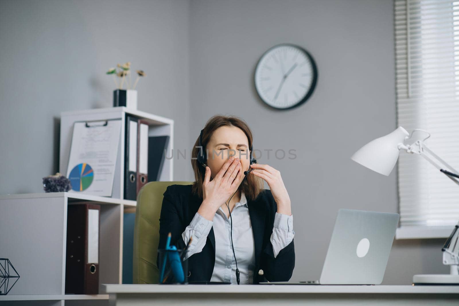 Tired woman in headphones sitting at the laptop computer while working in the office, then almost falling asleep and waking up. Indoor.