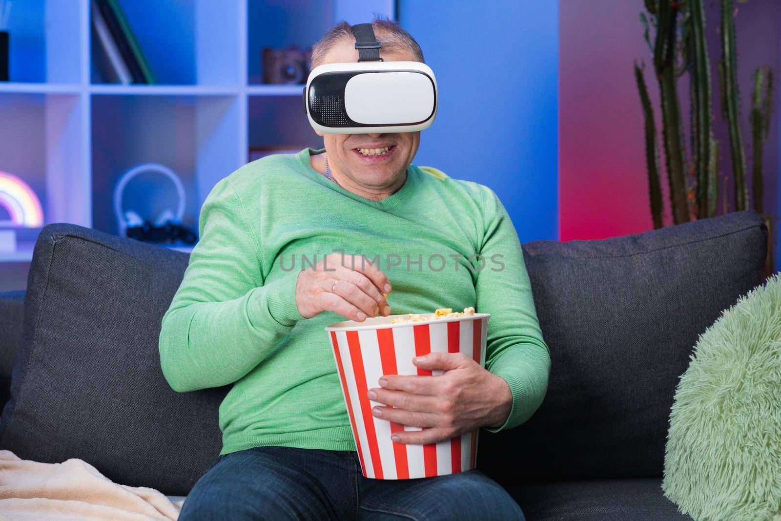 Senior man sitting on Sofa and holding popcorn, He wearing virtual reality glasses and lift up his hand to selective with VR Screen
