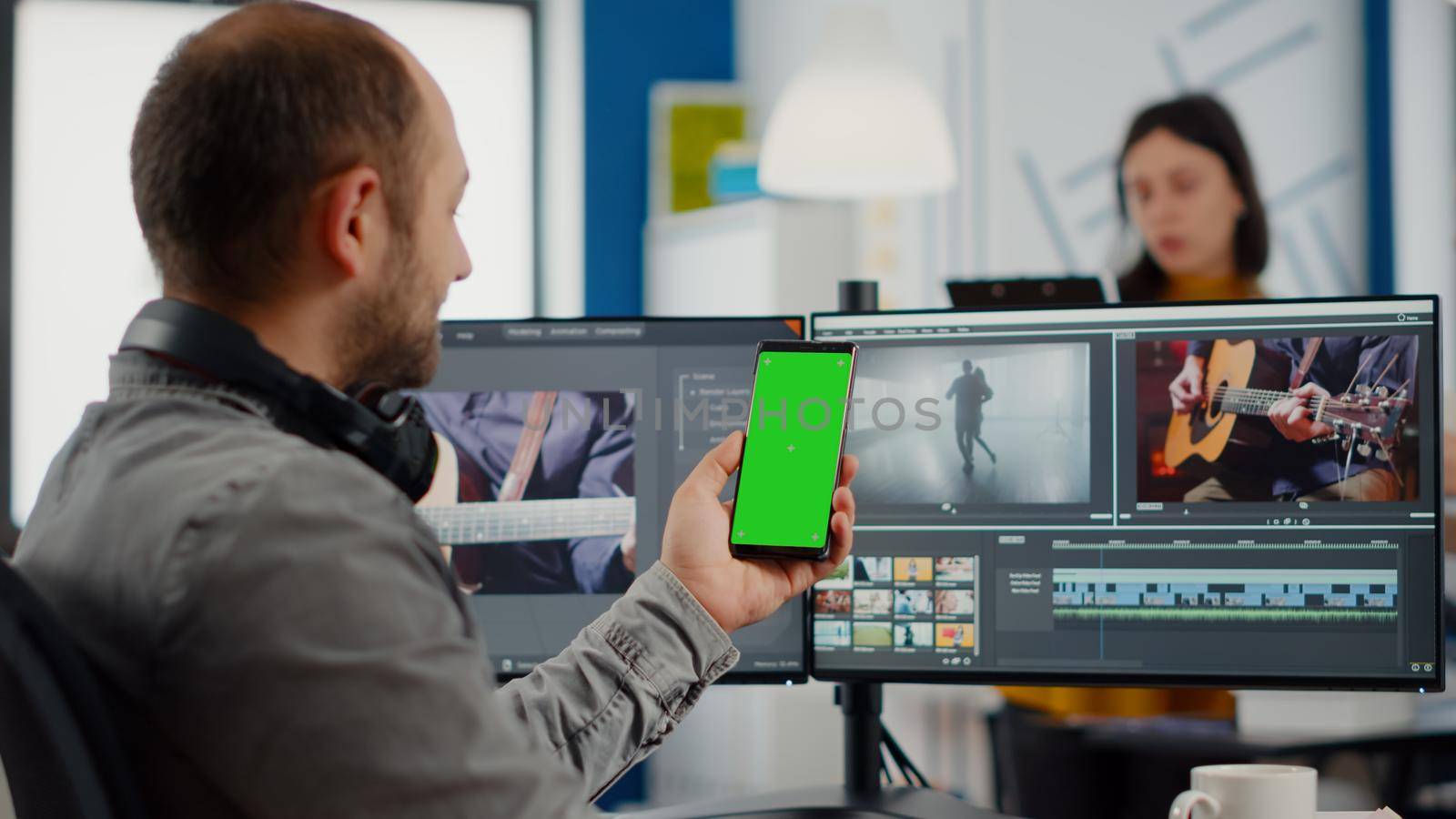 Video editor talking on video call holding smartphone with green screen, chroma key mock up isolated display taking break from retouching movie sitting in start up office. Team working in background