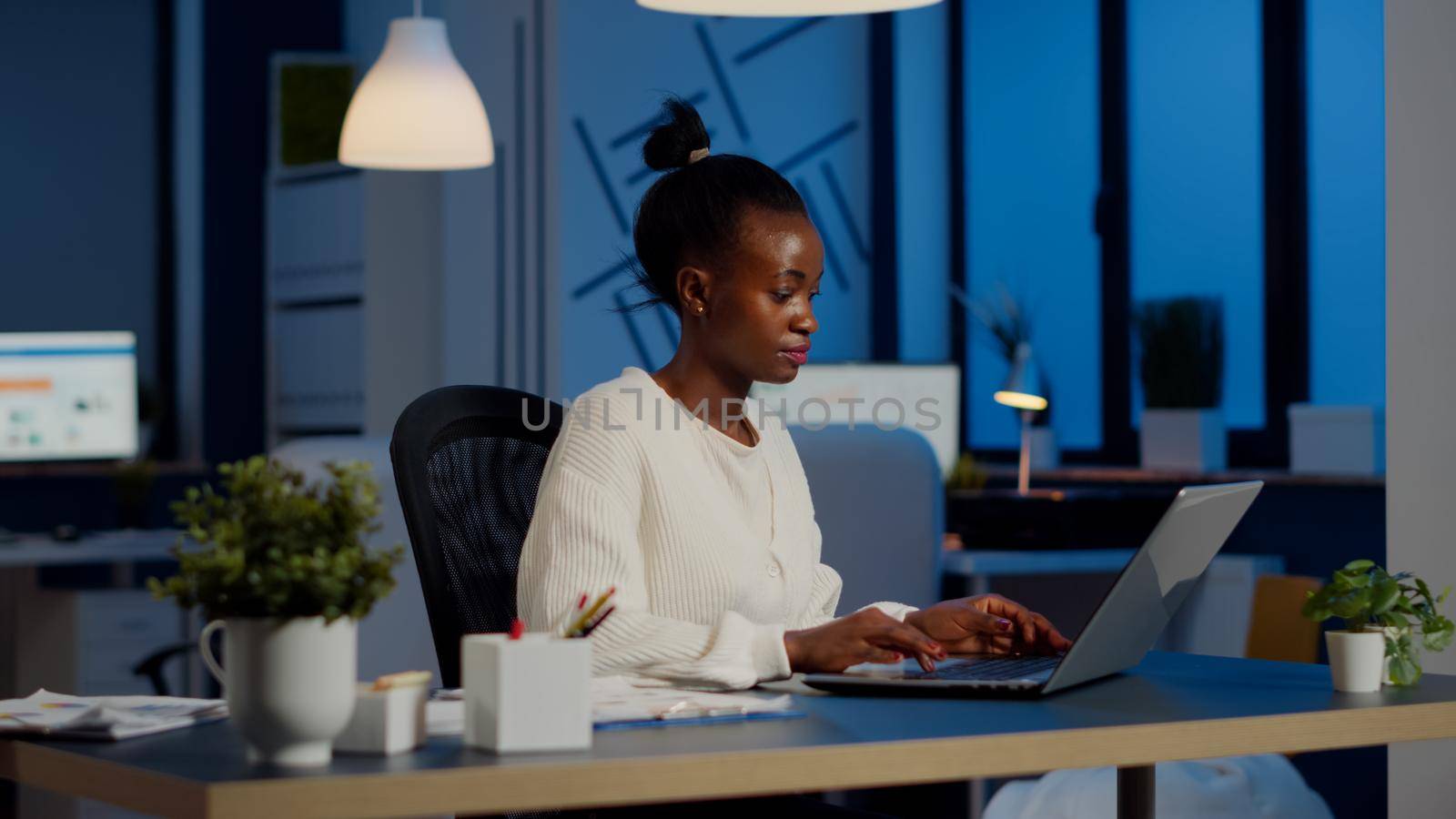 Busy african manager working on financial reports checking statistics graphs, typing on laptop sitting at desk late at night in start-up office doing overtime to respect deadline of financial project