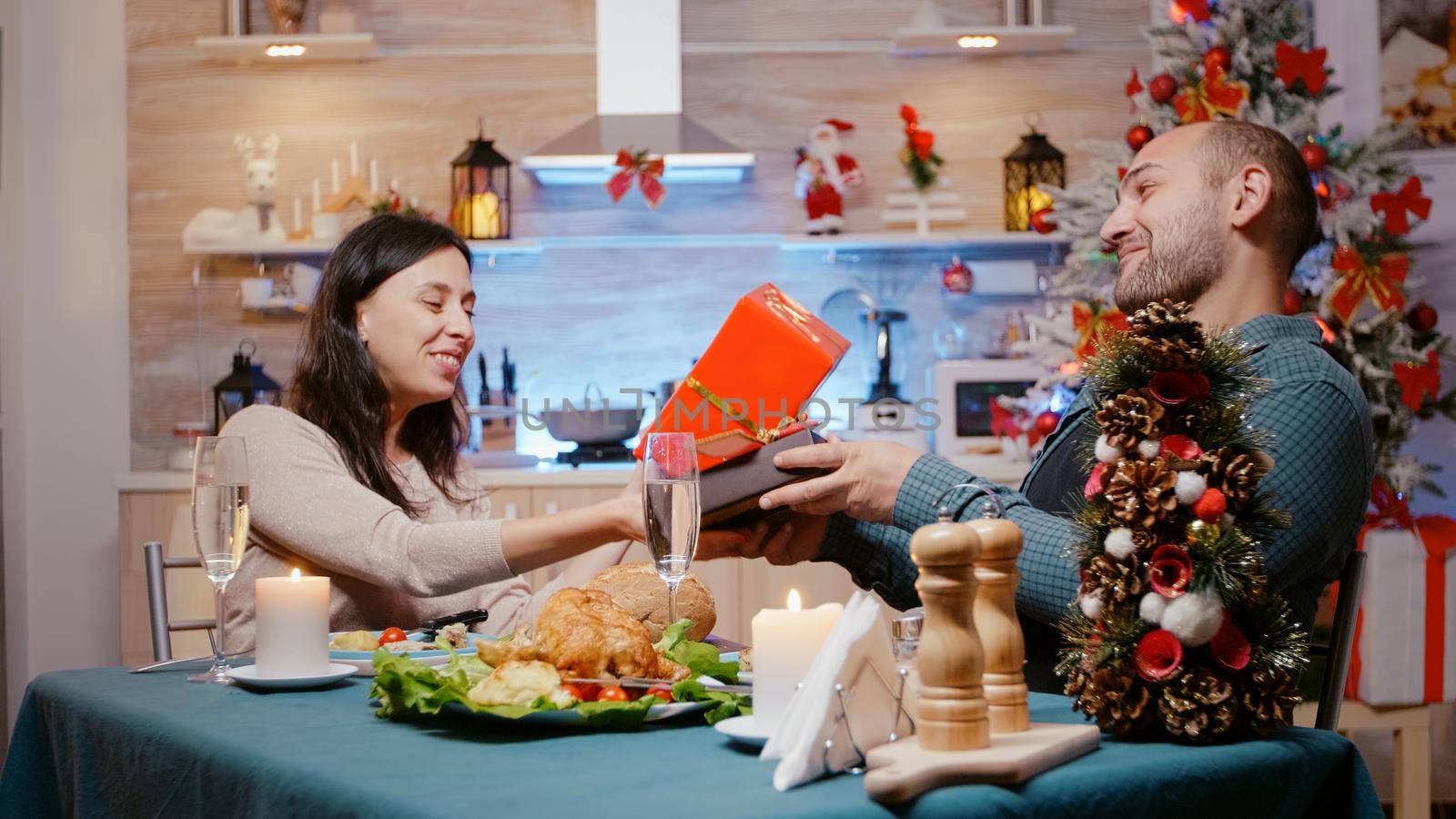 Festive couple exchanging gift boxes at christmas eve dinner. Man and woman celebrating traditional holiday while giving presents and enjoying festivity with food and glasses of champagne.