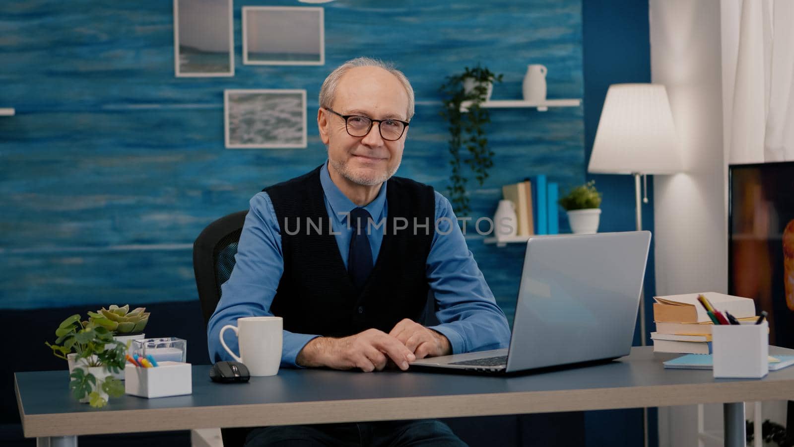 Senior man looking at camera smiling sitting in workplace at desk working from home while old wife watching tv in background. Retired business person prepared for online meeting using moden gadgets