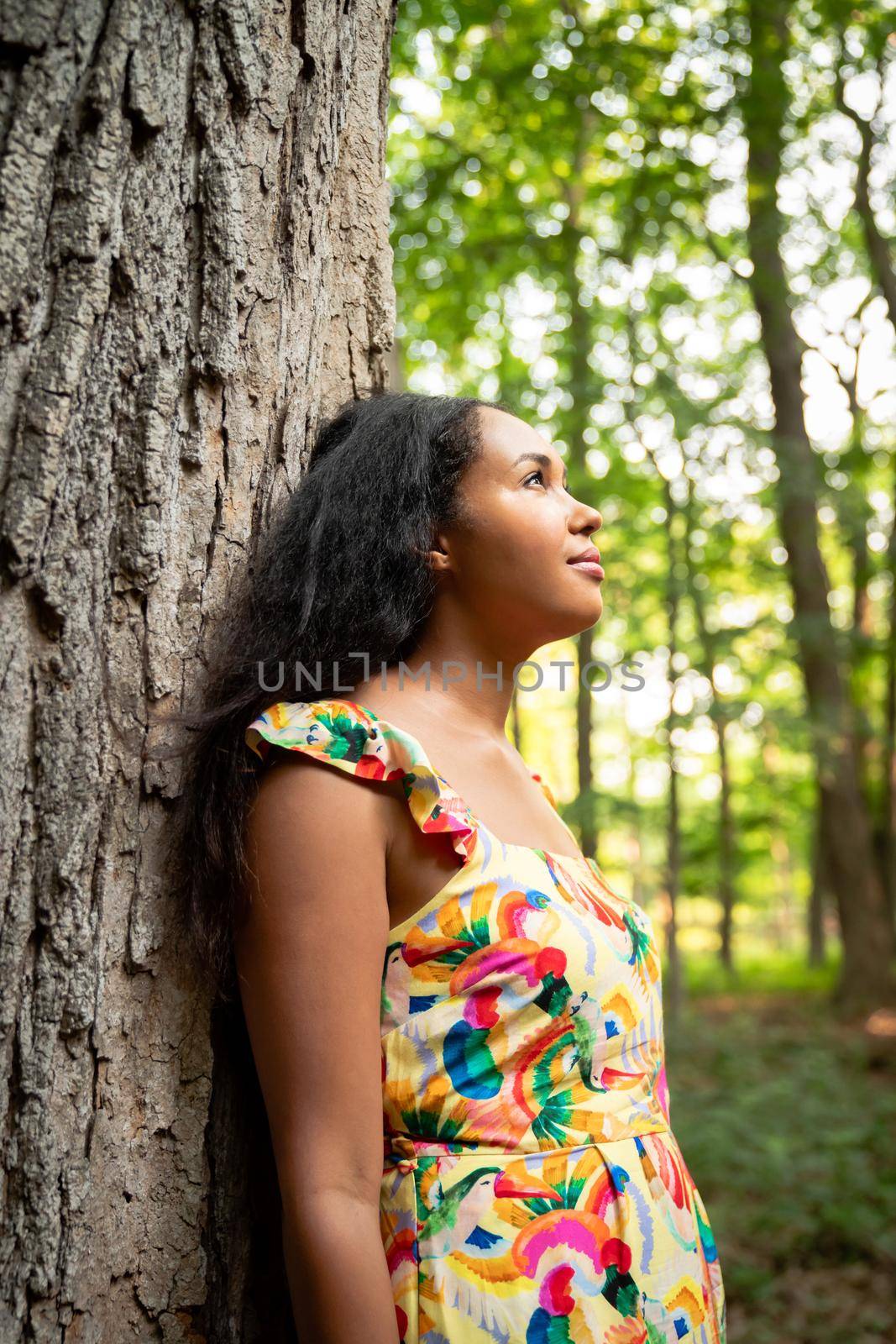 Beautiful portrait of a happy mixed race African American woman wearing a yellow floral dress leaning against a large rough bark covered tree trunk in a forest and looks up into the distance. by lapse_life