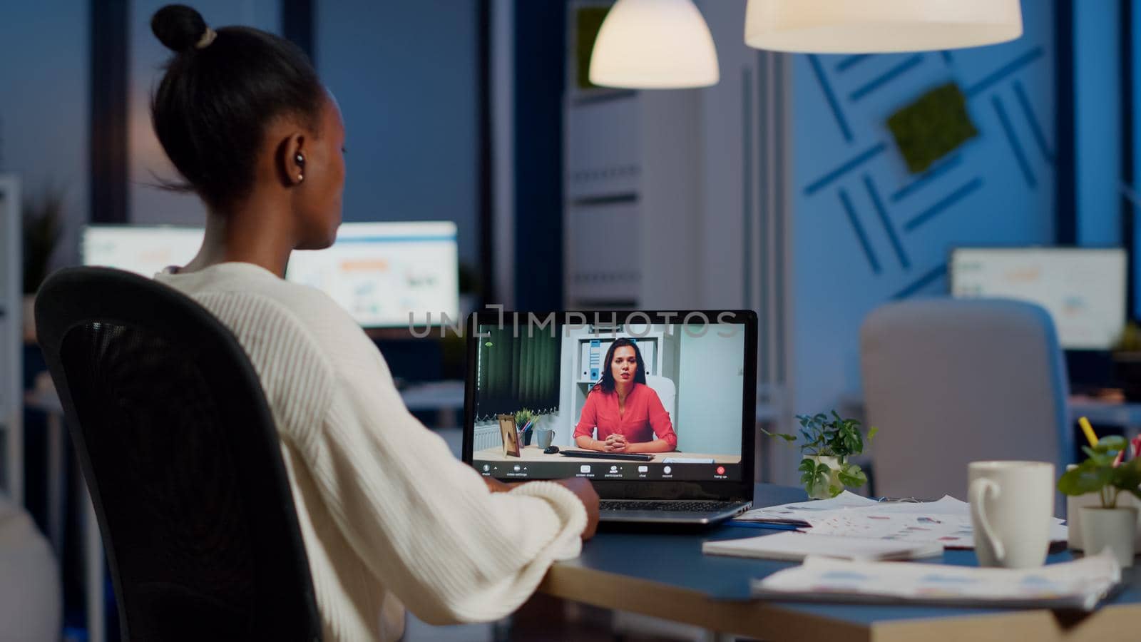 Dark skin manager woman talking with coworker during video conference at midnight from business office planning financial strategy. Worker using wireless headphones discussing at virtual meeting