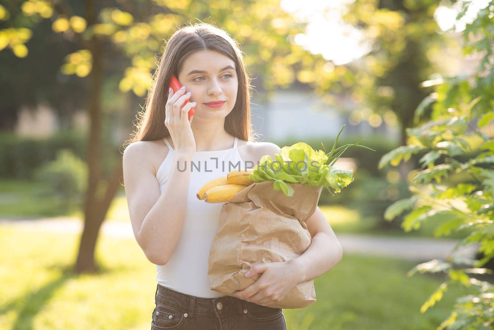 Happy girl student using a smart phone in a city park. European Girl Making Voice Message On Phone. Young brunette in a park. Woman speaks on the phone and smiles