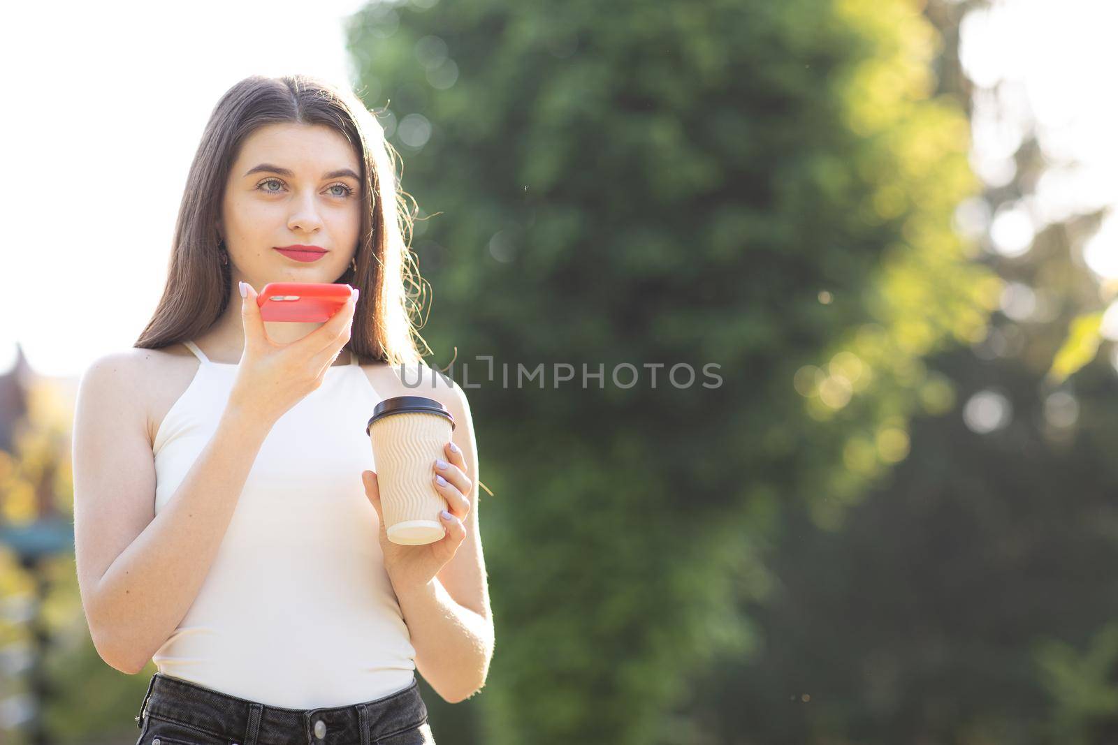 Young girl walking in the Park speaking on Smartphone. Happy girl using a smart phone voice recognition audio ai message speech function on line walking in park. Having cup with Coffee in her Hands