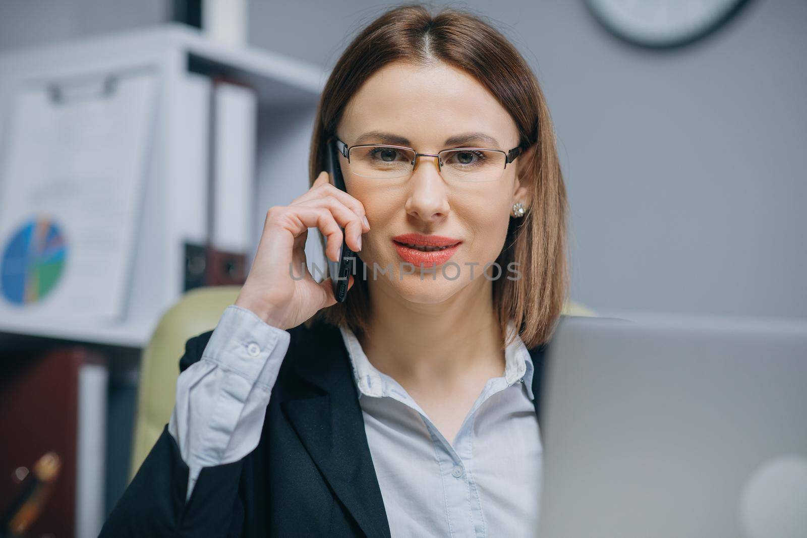 A beautiful smiling young businesswoman wearing glasses is using her smartphone while sitting at the desk with a laptop in the office indoors