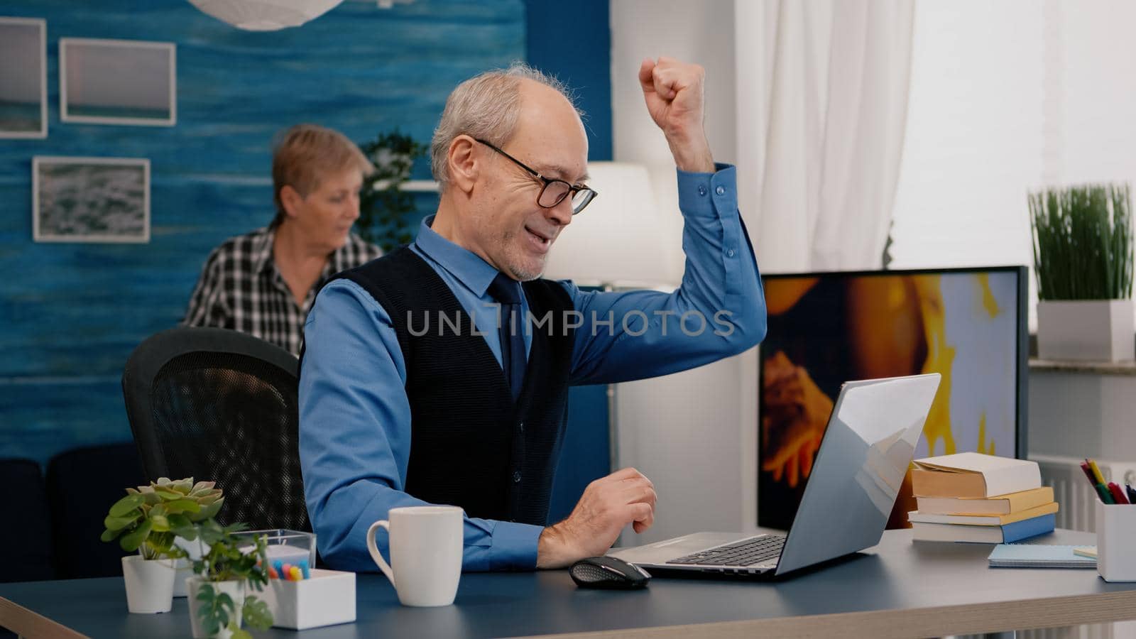 Excited senior man feel ecstatic reading great online news on laptop working from home. Retired employee using modern technology reading typing, searching while senior wife sitting on couch