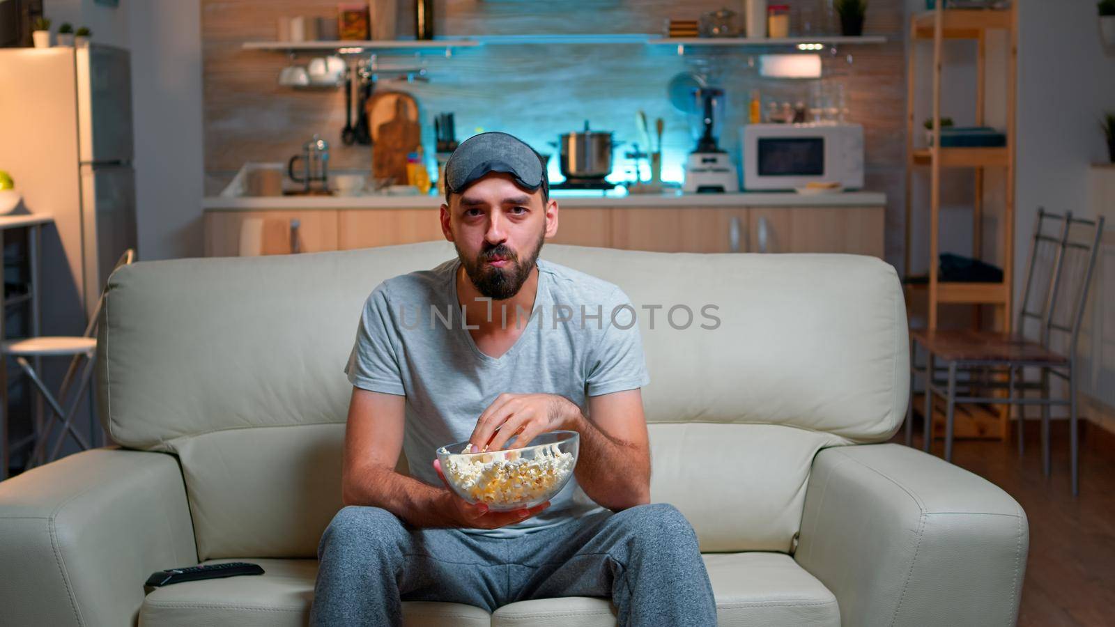 Portrait of man with beard holding popcorn bowl while watching entertainment movie on television. Concentrated person with eye sleep mask sitting on sofa late at night in kitchen