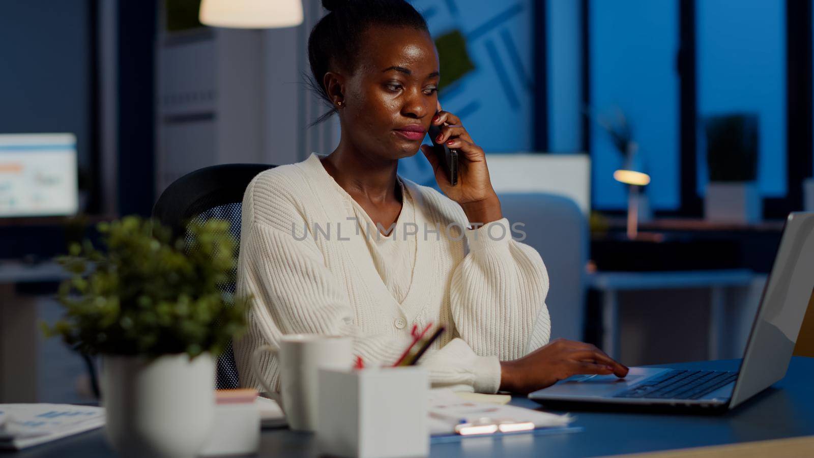 African employee speaking at phone while working at laptop late at night by DCStudio