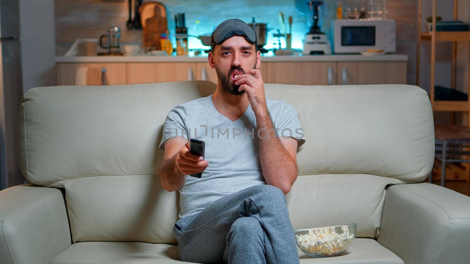 Adult man eating popcorn while standing in front of television on sofa looking at tv lifestyle shows. Caucasian male with eye sleep mask relaxing alone late at night in kitchen