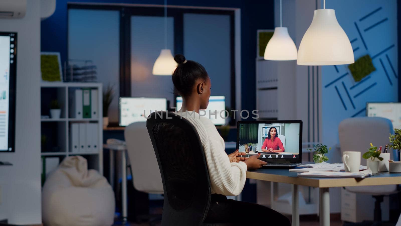 African freelancer working remotely discussing with woman partner online sitting in front of laptop in start-up office talking during virtual meeting at midnight, using headphone doing overtime