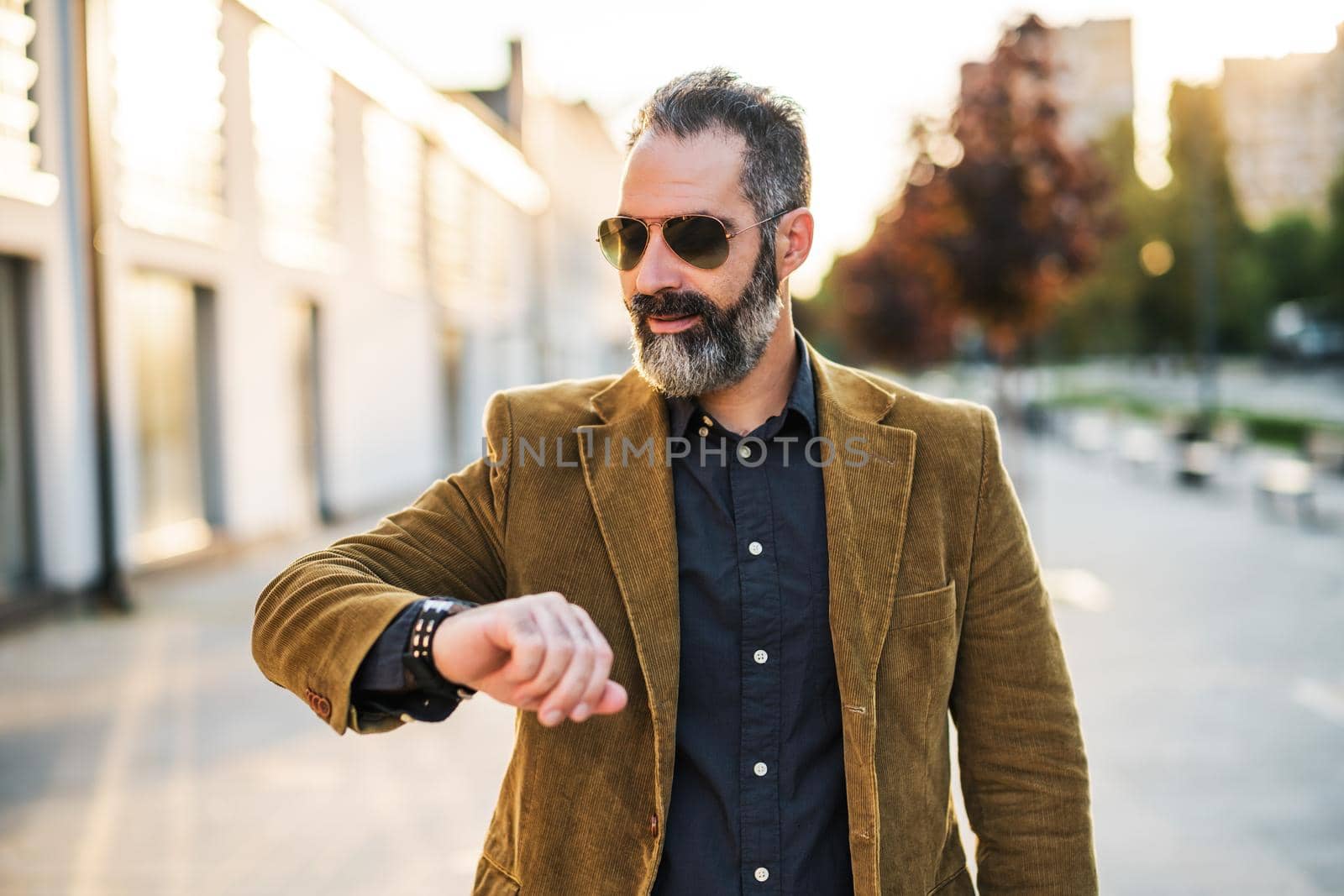 Modern businessman with beard looking at watch and walking on the city street.