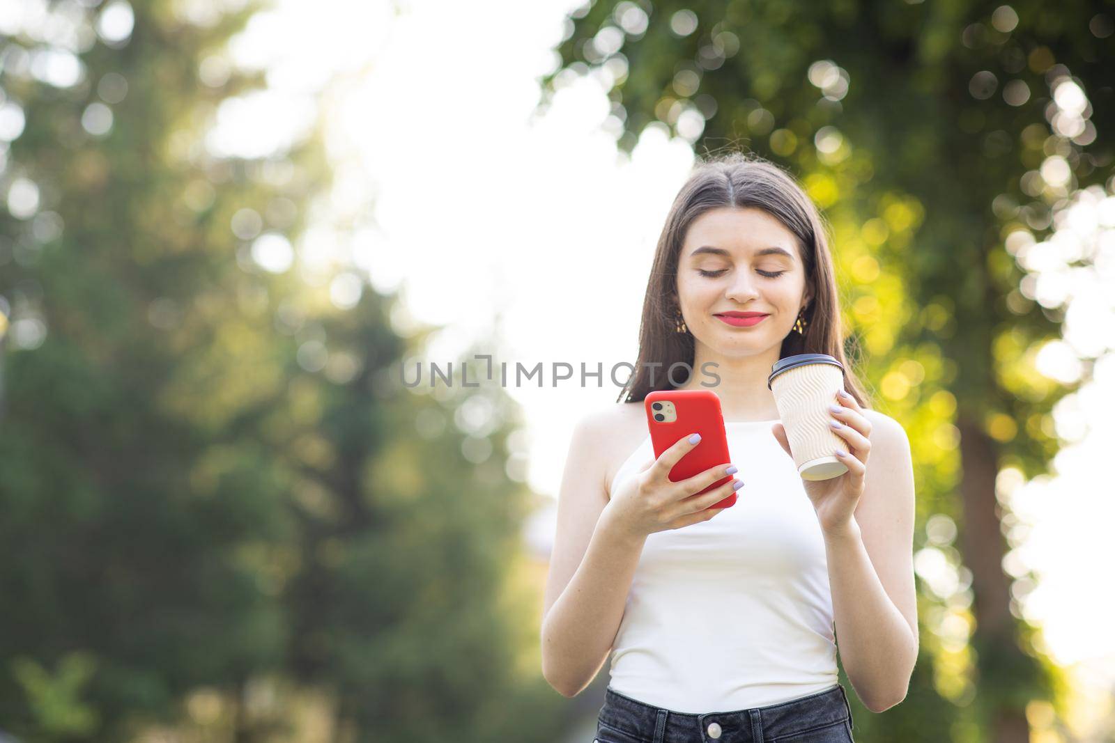 Girl Laughing while looking on her Smartphone. Town Landscape with trees. Cheerful young girl with a paper Cup of coffee in her left hand.