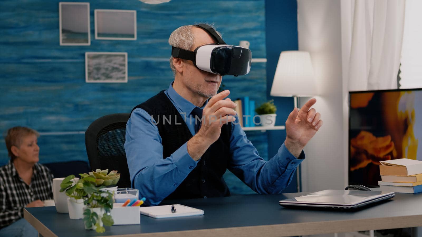 Retired man experiencing virtual reality using vr headset in living room working from home. Old remote employee searching, analysing financial reports while elderly wife watching tv in background