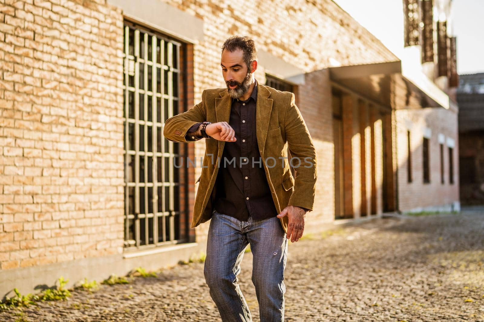 Businessman with beard in panic looking at watch and walking in hurry on the city street.