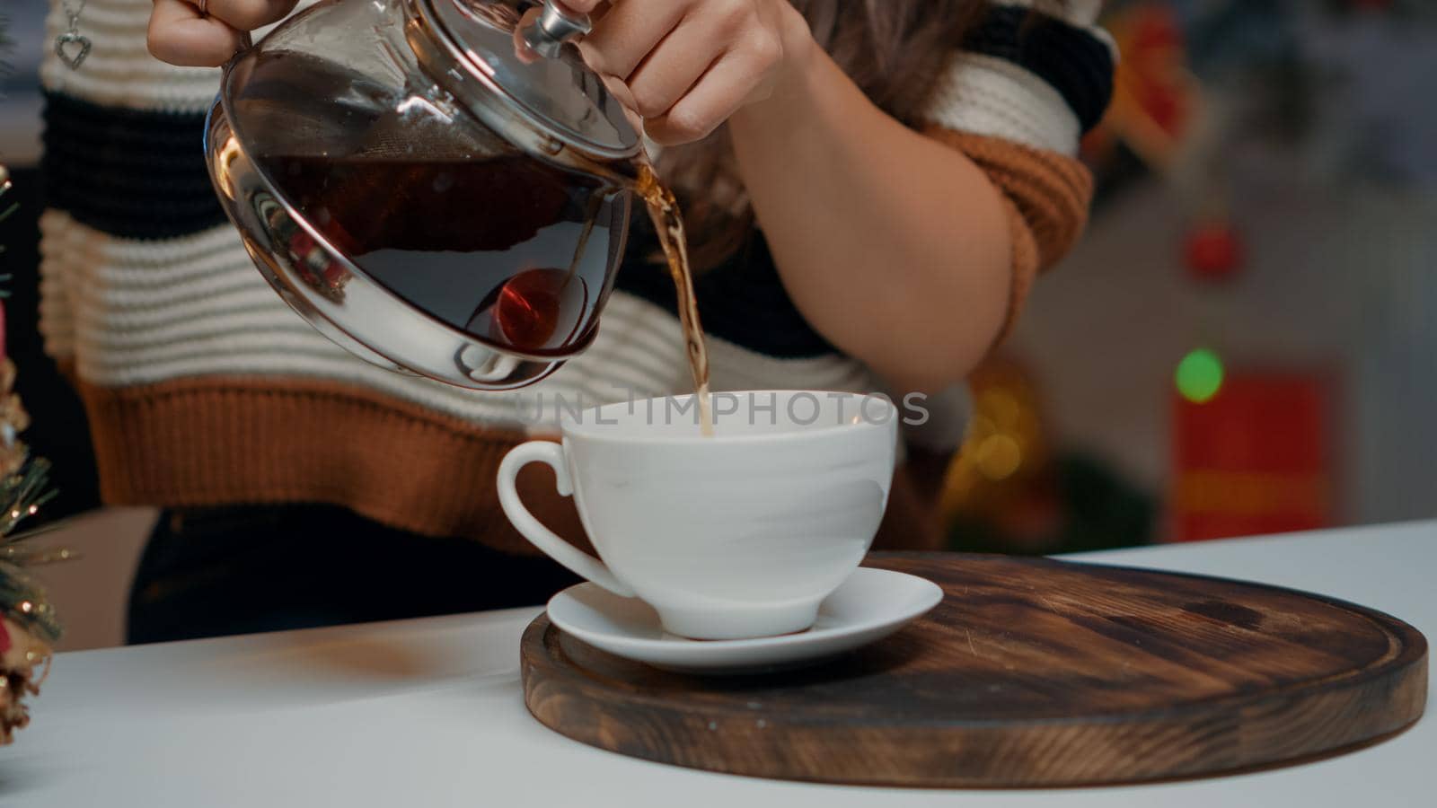 Close up of pouring beverage from tea kettle at festive home. Happy woman preparing for christmas dinner with hot drink for warmth in december. Kitchen with decorations, ornaments and gifts