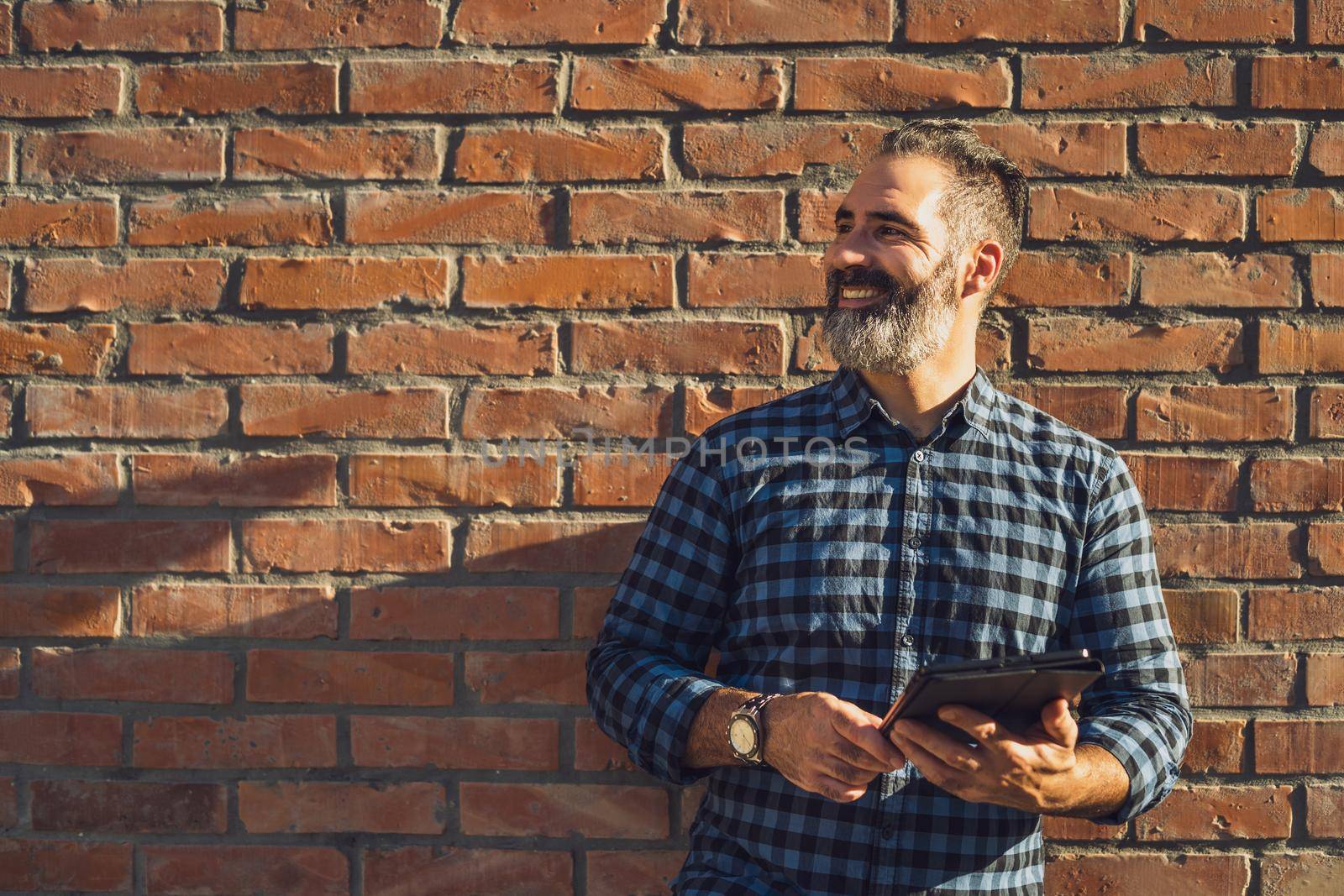 Portrait of modern businessman with beard using digital tablet while standing in front of brick wall outdoor.