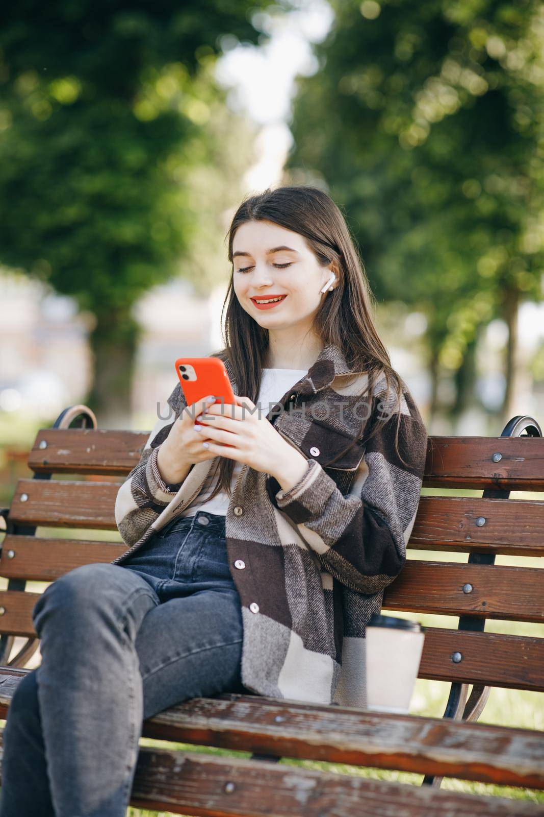 Girl wearing earphones and using smartphone listening music. Attractive woman browsing on mobile phone in park. City, urban background. by uflypro