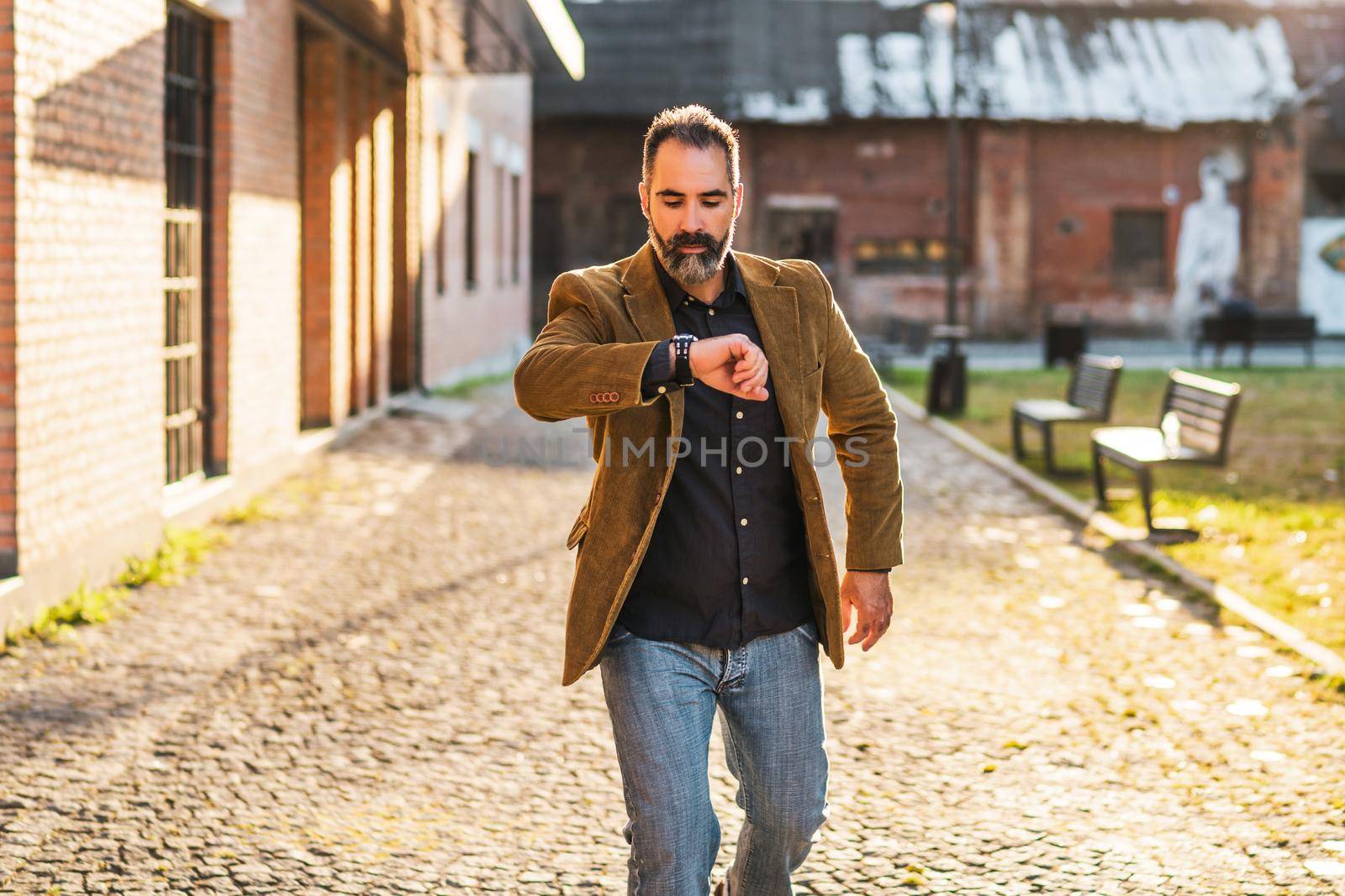 Modern businessman with beard looking at watch and walking in hurry on the street.