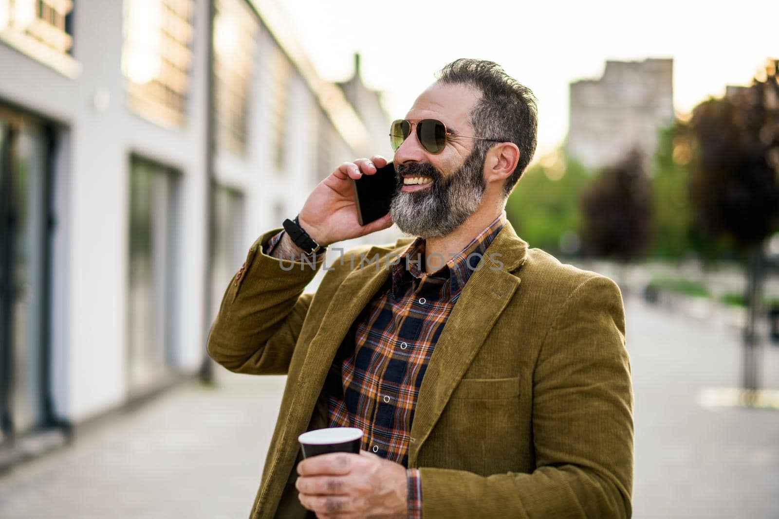 Portrait of modern businessman with beard using mobile phone and drinking coffee while standing on the city street.