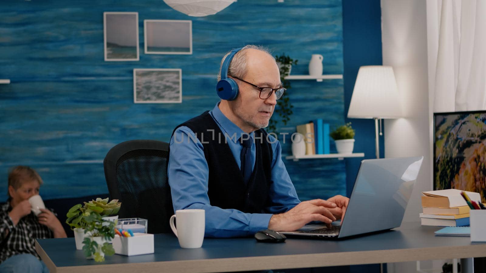 Focused old entrepreneur with headphones listening music while checking graphics writing on laptop working from home. Retired man using modern technology reading typing, searching on computer