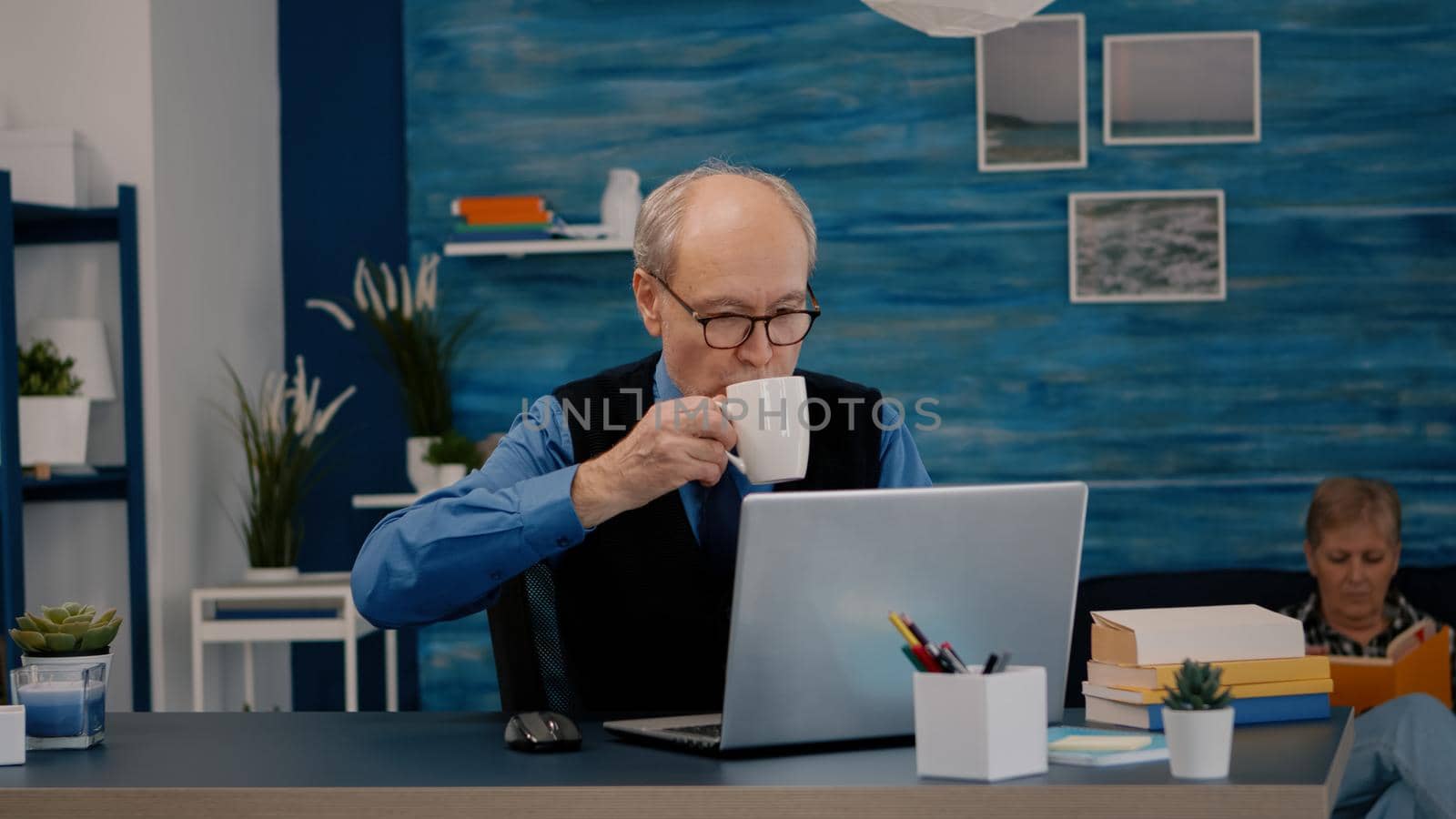 Senior businessman reading reports sitting in front of laptop working from home drinking coffee. Retired man using modern technology analysing typing searching while wife sitting on couch reading a book