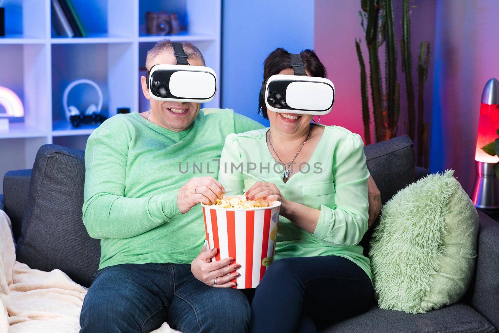 Family couple is sitting at the sofa with popcorn and watching TV using VR glasses. Old Caucasian Spouses Sitting Together at Sofa Eating Popcorn and Watching Movie in VR Goggles.