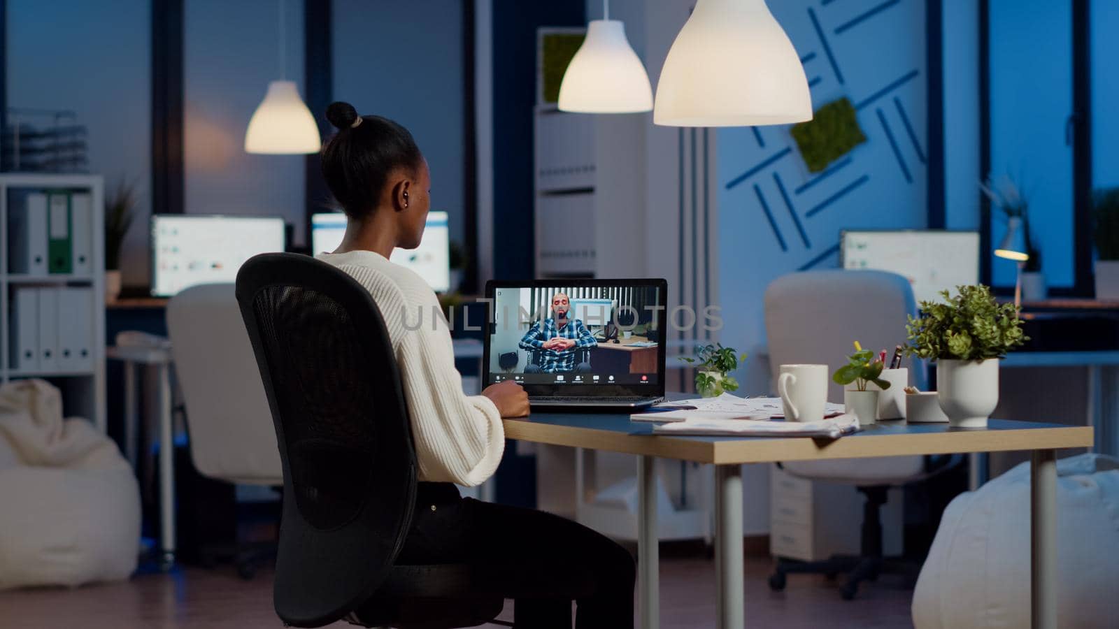 Black woman freelancer discussing with paralyzed client on video call at midnight from business office using headphone. Businesswoman using virtual conference talking on webcam during online meeting