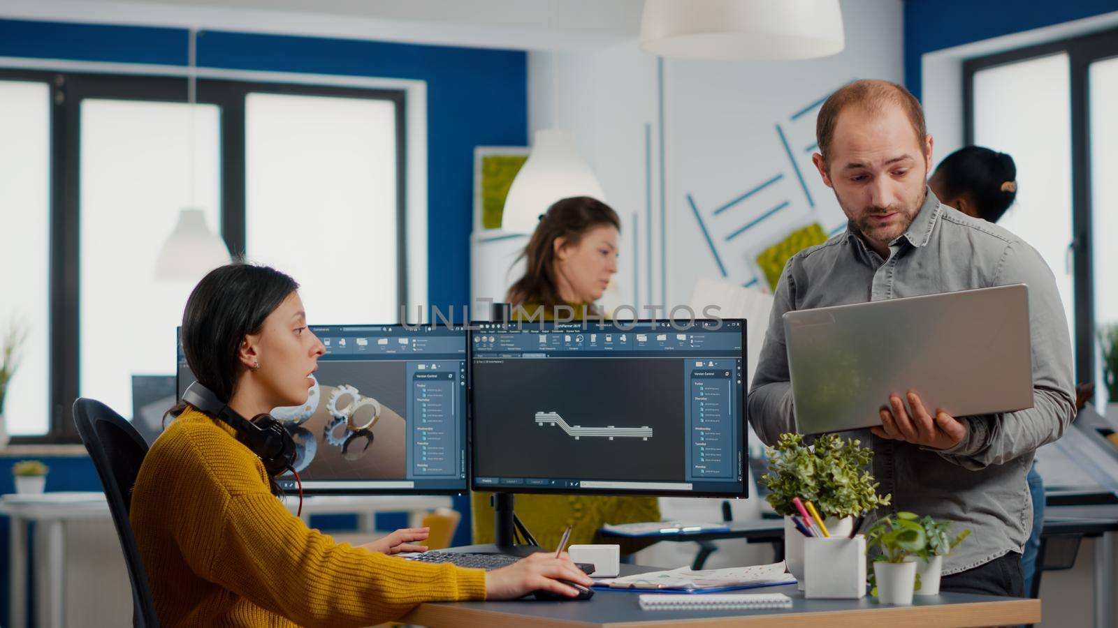 Man project supervisor holding laptop and talking with woman industrial engineer who works on computer the development of new machinery prototype. Diverse team working in creative agency office