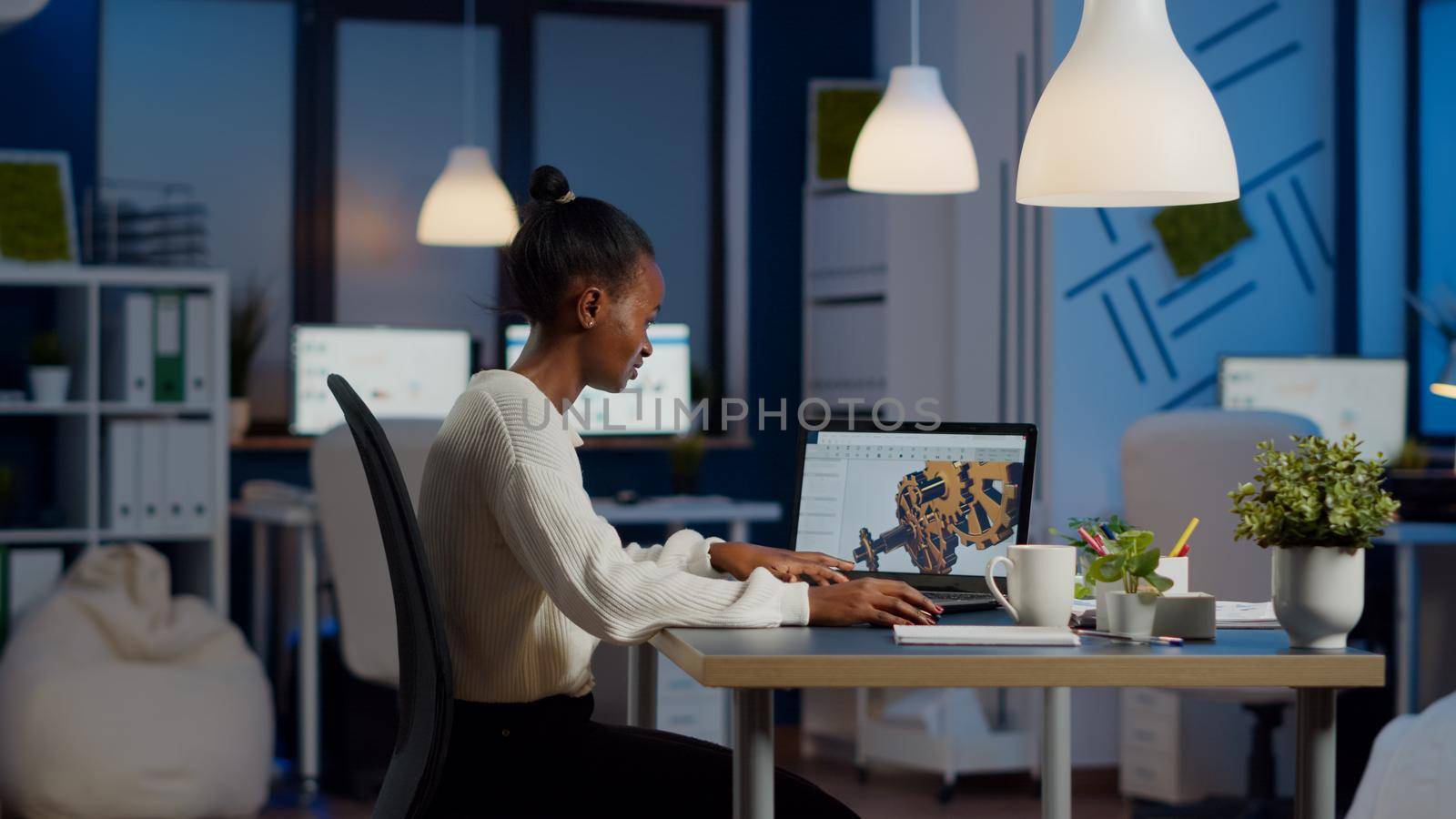 African engineer working late at night on a 3D model of industrial gears at laptop doing overtime in start up office. Freelancer studying prototype idea on pc showing cad software on device display