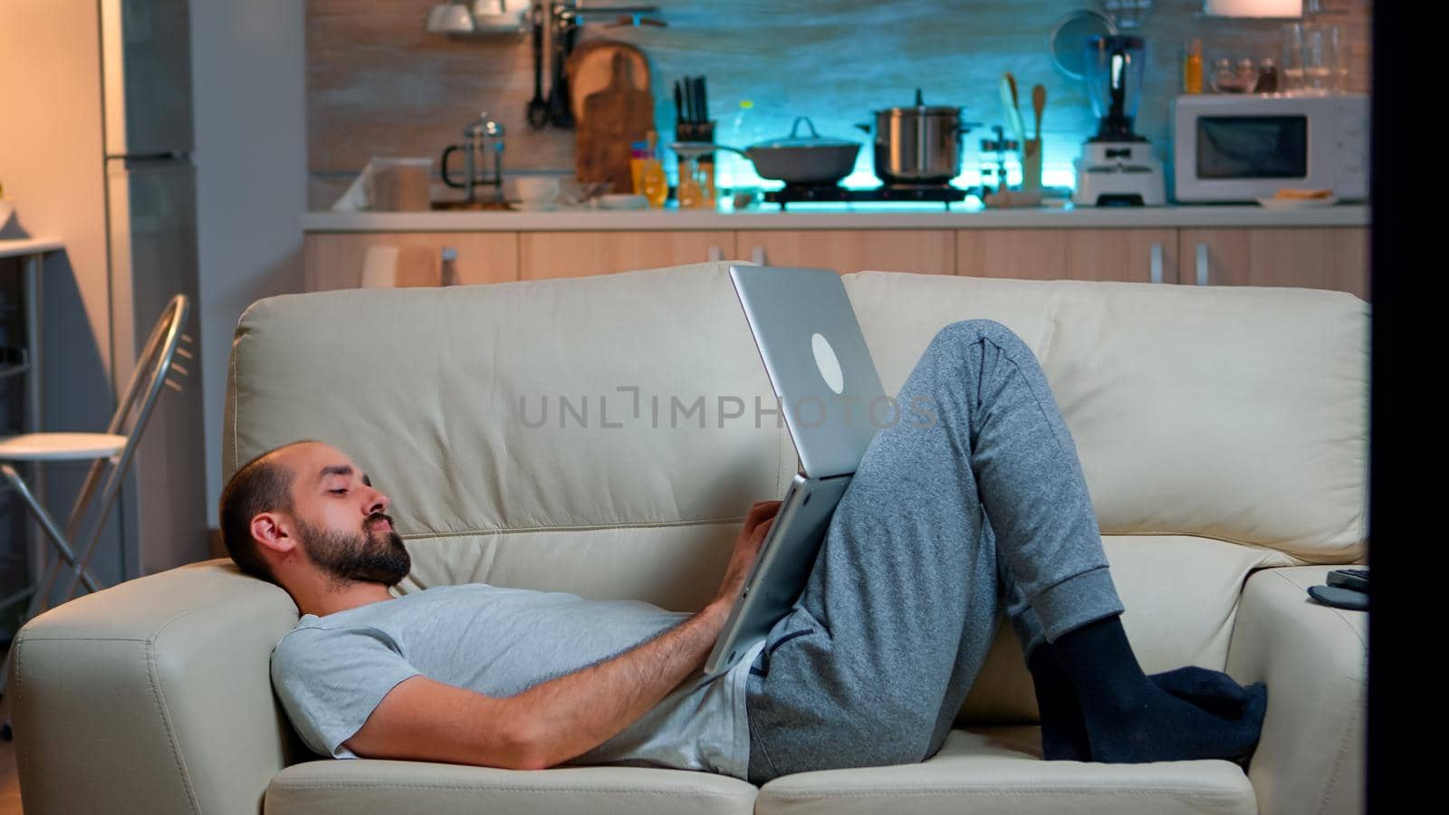 Man falling asleep in fron of TV while working on the laptop. Cozy man lying down on sofa while typing on laptop computer for social media project. Exhausted man working