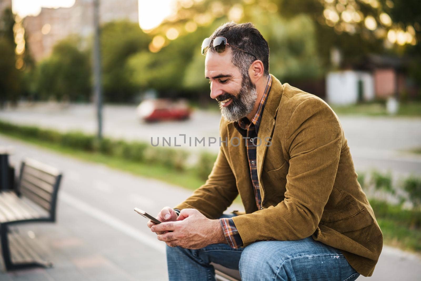 Modern businessman with beard using mobile phone while sitting on the bench in the city street.