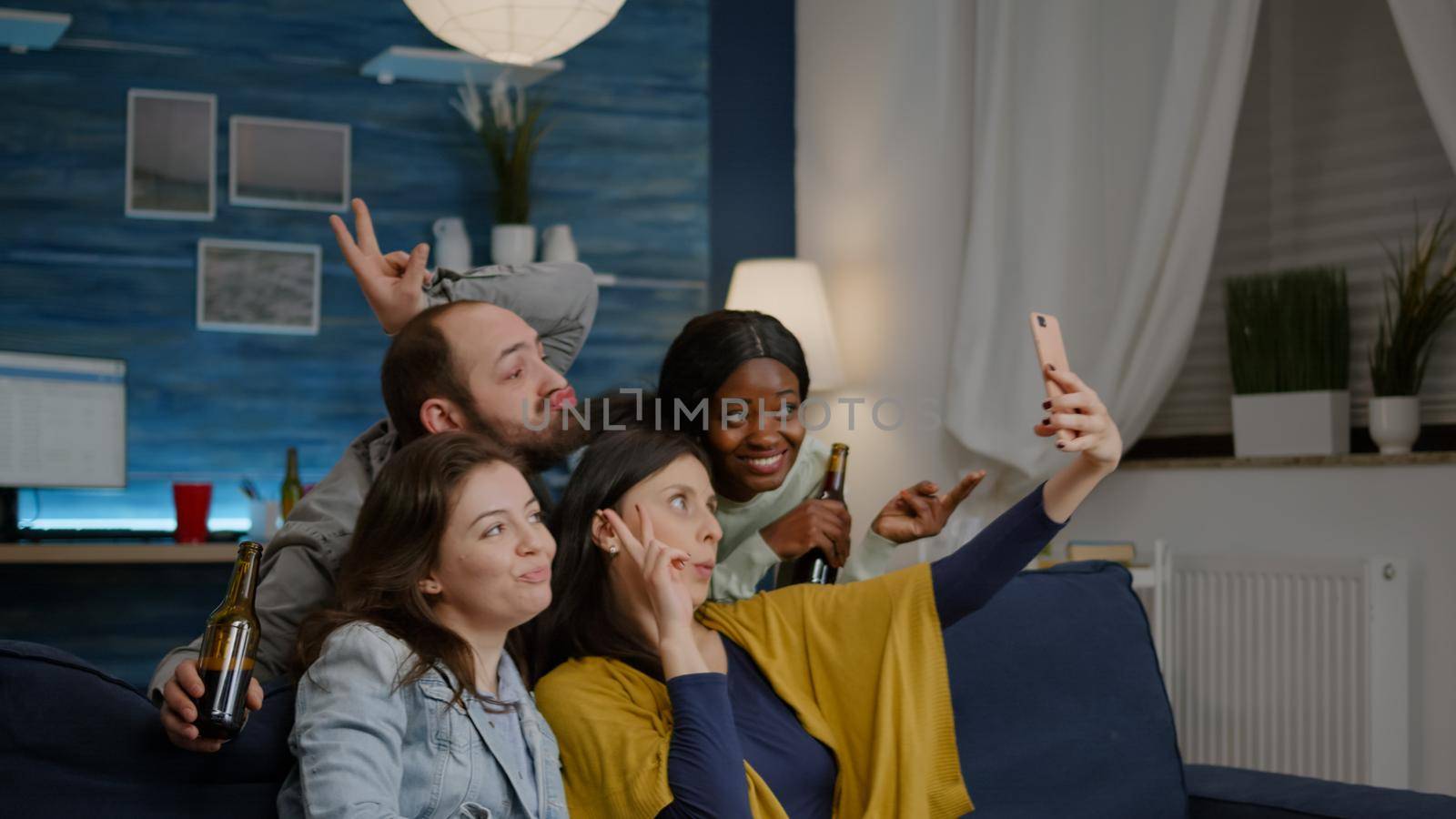 Multiracial friends resting on sofa enjoy spending time together while taking selfie sharing on social media. Group of multi-ethnic people laughing late at night in living room