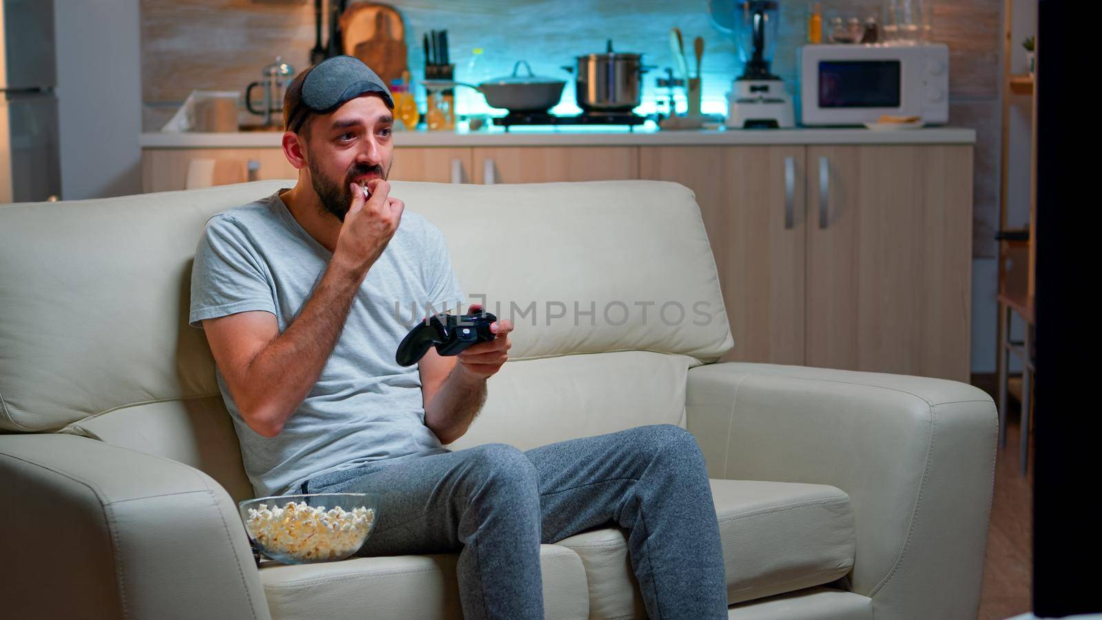 Upset pro gamer sitting on couch and playing soccer videogames for online competition using gaming joystick. Disappointed man looking at television eating popcorn late at night in kitchen